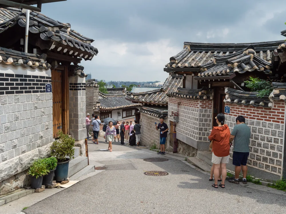 A sloping alley in Bukchon lined with tiled hanok roofs on both sides, the modern city skyline visible in the gap beyond