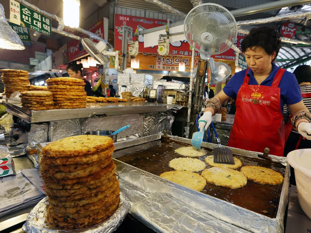 A vendor frying golden bindaetteok mung-bean pancakes on a sizzling griddle beside tall stacks of finished pancakes at a Gwangjang Market stall
