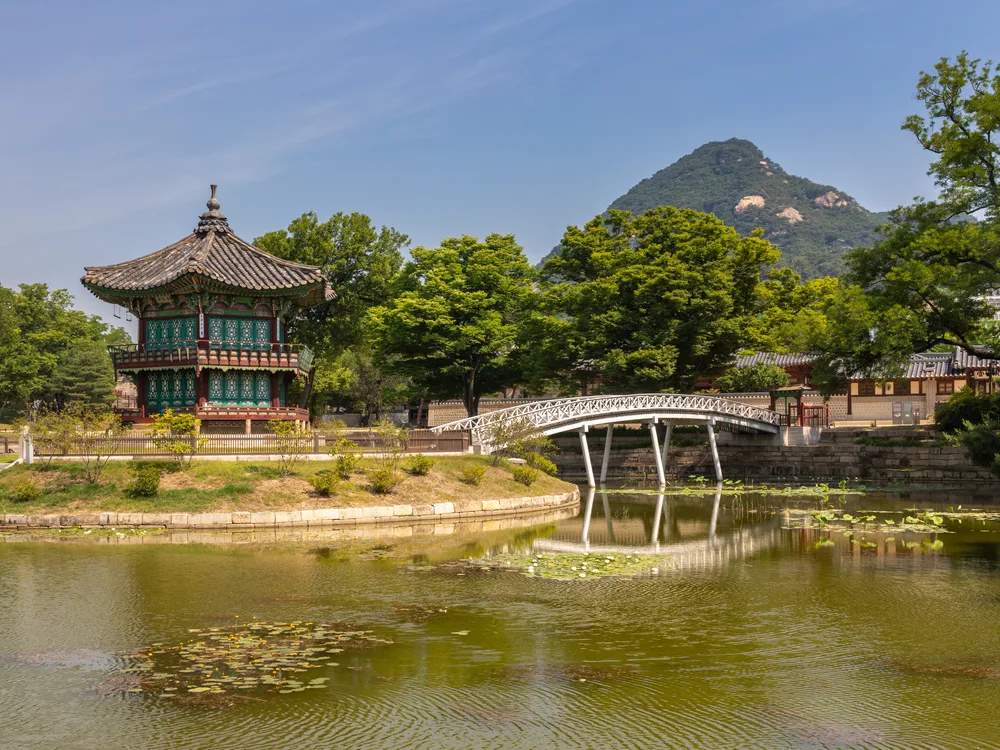 The hexagonal Hyangwonjeong pavilion on its island in a lily-covered pond at Gyeongbokgung, a white footbridge and a mountain behind