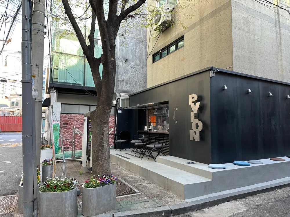 A small black-clad corner cafe in Seongsu-dong with an open serving window, bar stools and flower planters, wedged against an older building