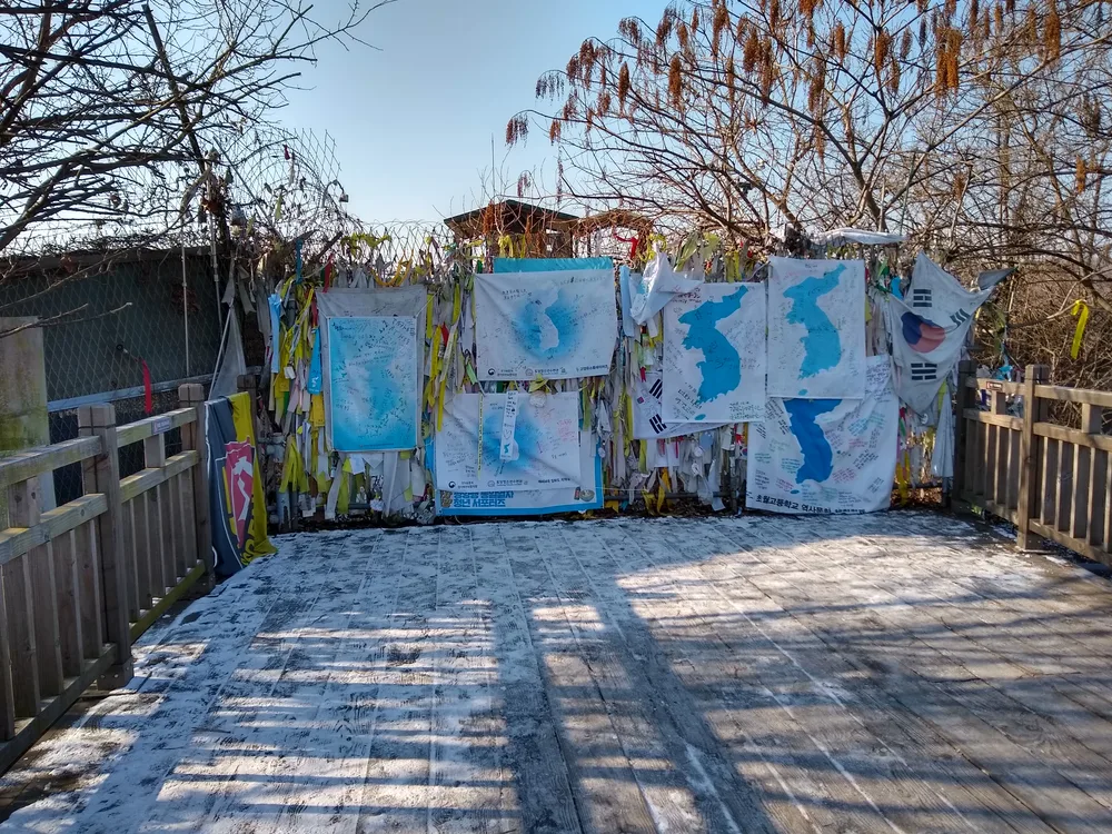 A wire fence at Imjingak covered in coloured prayer ribbons and hand-painted maps of the Korean peninsula, with snow on the boardwalk