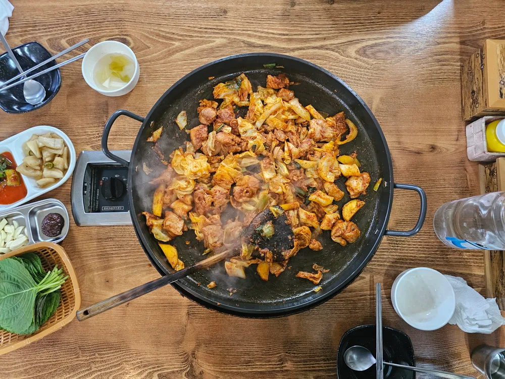 A large pan of spicy stir-fried dak-galbi chicken with cabbage, cooking on a table burner with side dishes