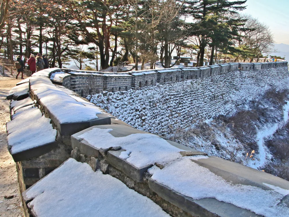The Namhansanseong fortress wall snaking along a snow-covered ridge, hikers in winter gear walking the path beside it