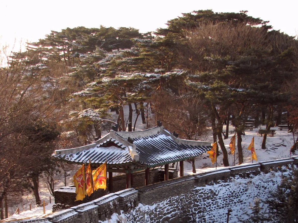 A snow-covered gate and tiled pavilion at Namhansanseong with yellow military flags, a snowy path leading to an arched gate
