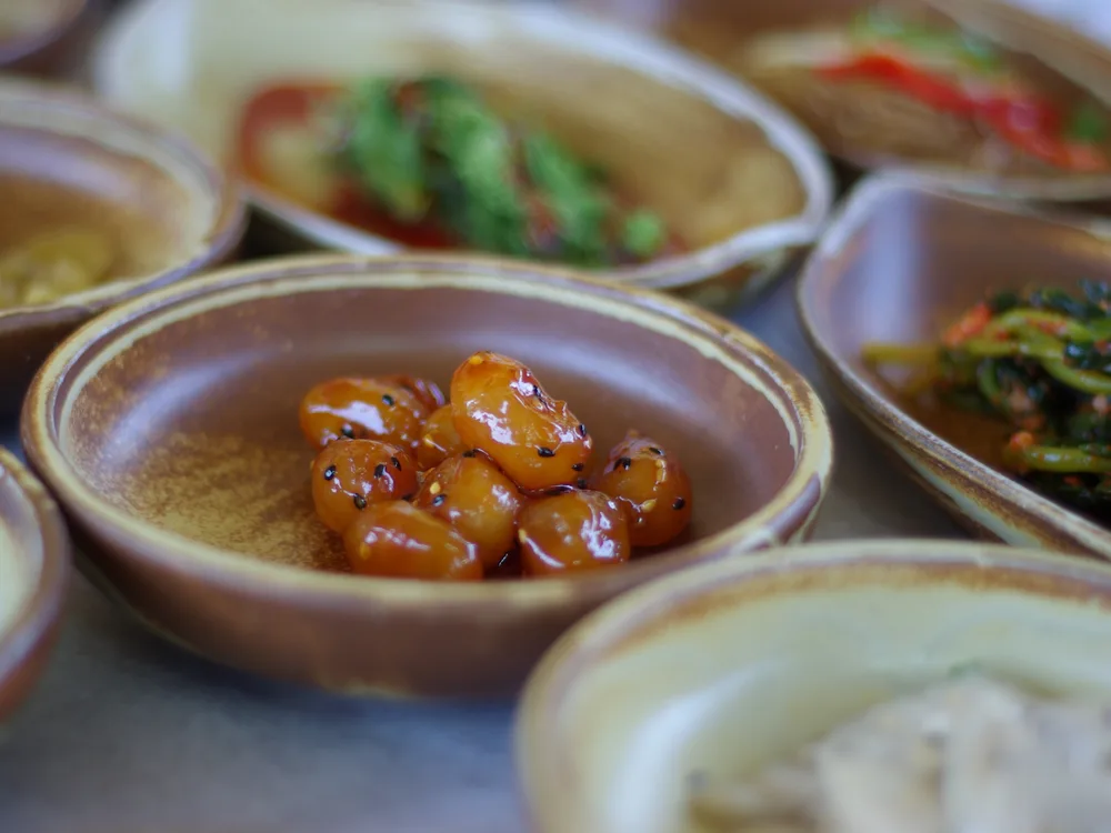A close-up of small brown ceramic bowls of Korean side dishes, one holding glossy soy-glazed beans