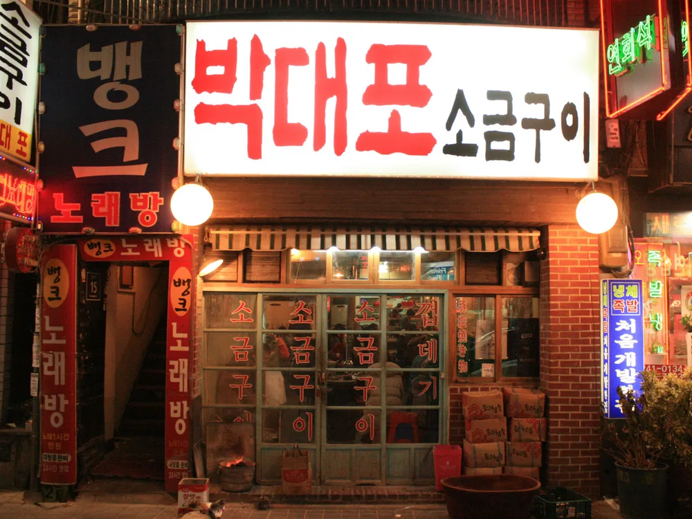 The lit-up front of a neighbourhood samgyeopsal grill house in Korea at night, with a hand-painted red sign and diners visible through the window