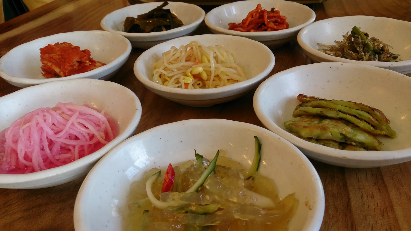 Eight white dishes of Korean banchan — kimchi, seasoned bean sprouts, pink pickled radish, a savoury pancake, acorn jelly — spread across a wooden restaurant table