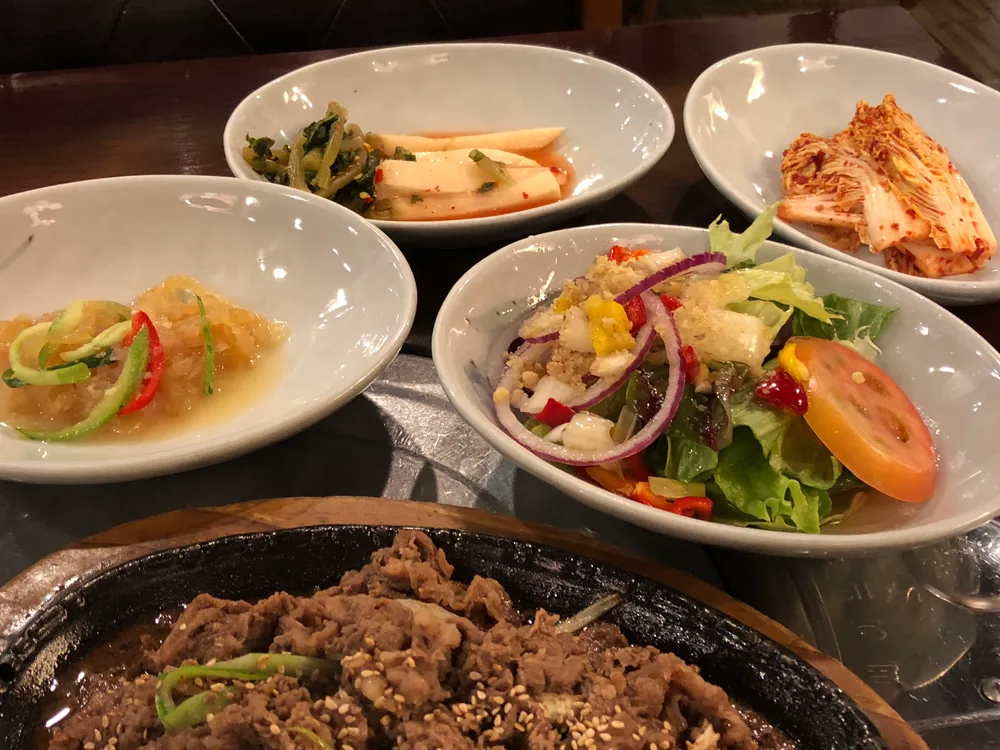 Four small white dishes of Korean side dishes — kimchi, pickled radish, a jellyfish salad, and a green salad — around a sizzling plate of marinated beef