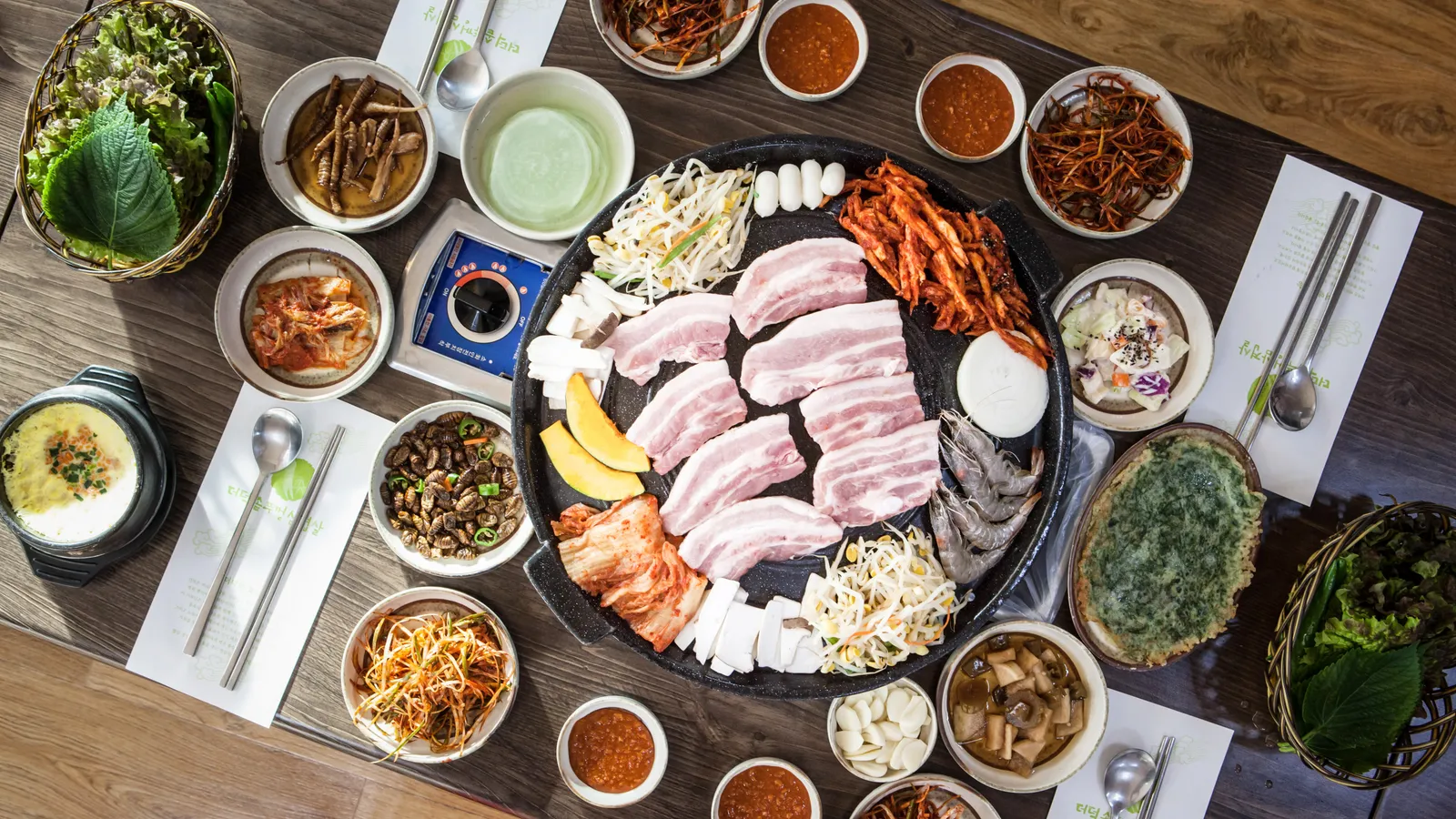 A top-down view of a samgyeopsal table — raw pork belly on a round grill plate ringed by bean sprouts and kimchi, with baskets of lettuce and a dozen small side dishes