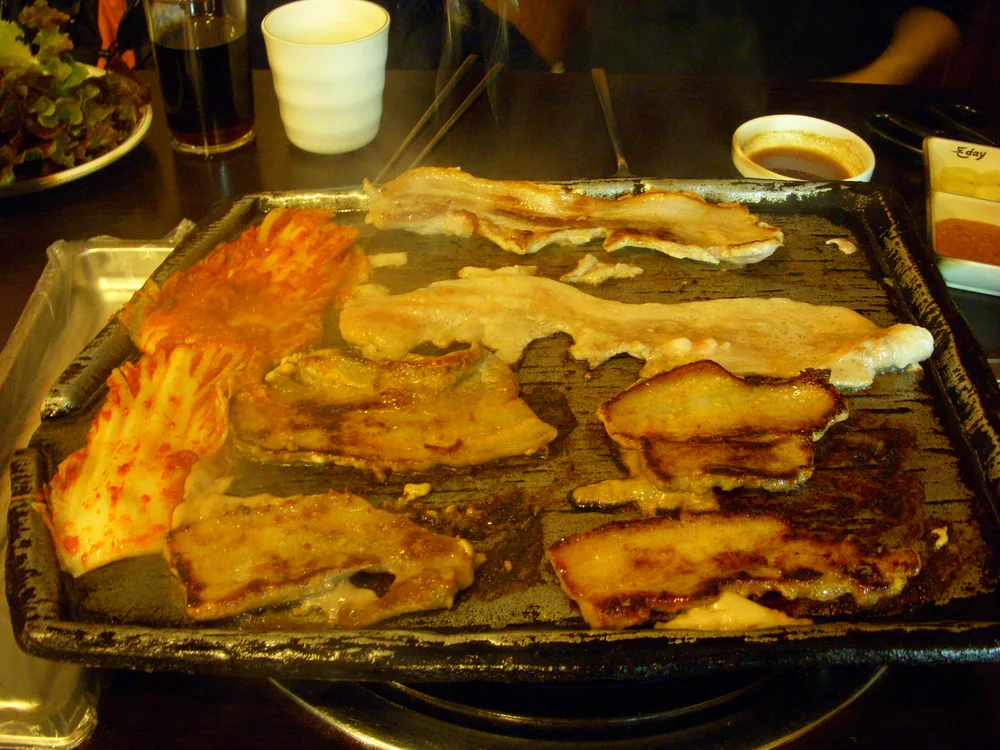 Strips of pork belly and a wedge of kimchi cooking on a dark cast-iron grill plate, a small dish of dipping sauce beside it