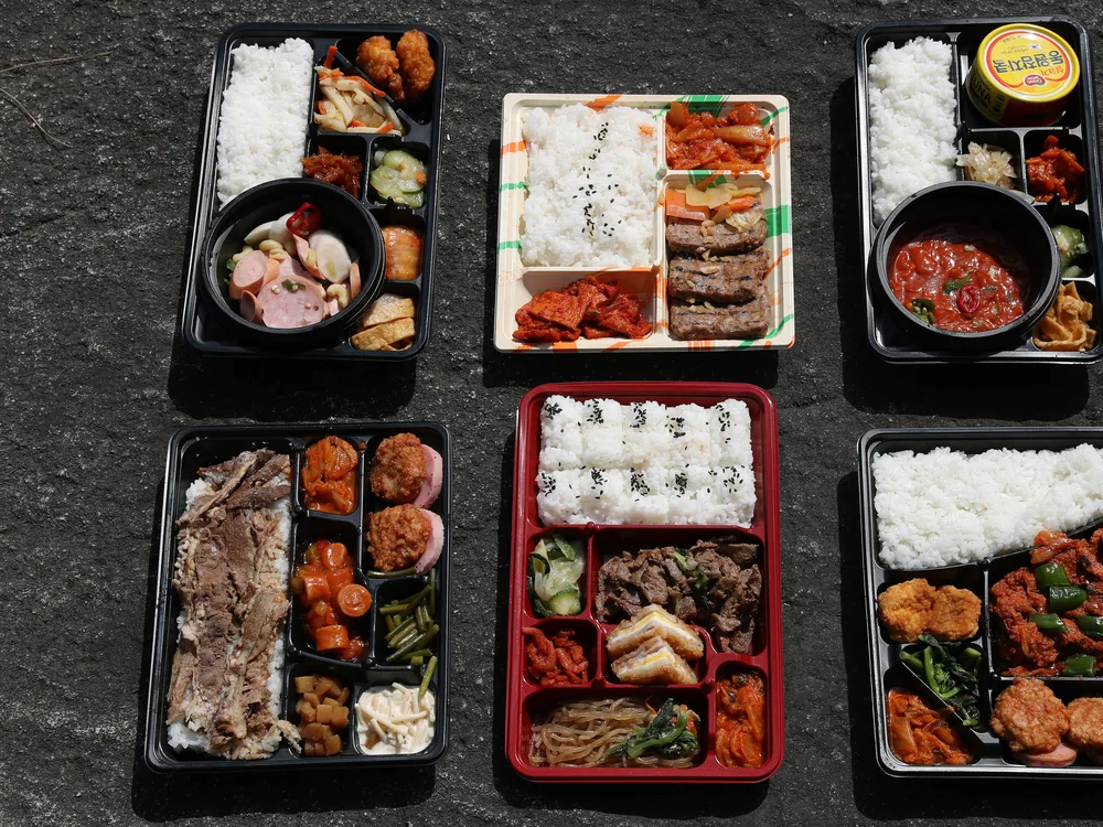Six convenience-store lunchboxes laid out, each with a block of rice and compartments of meat, sausage, egg, kimchi and other side dishes