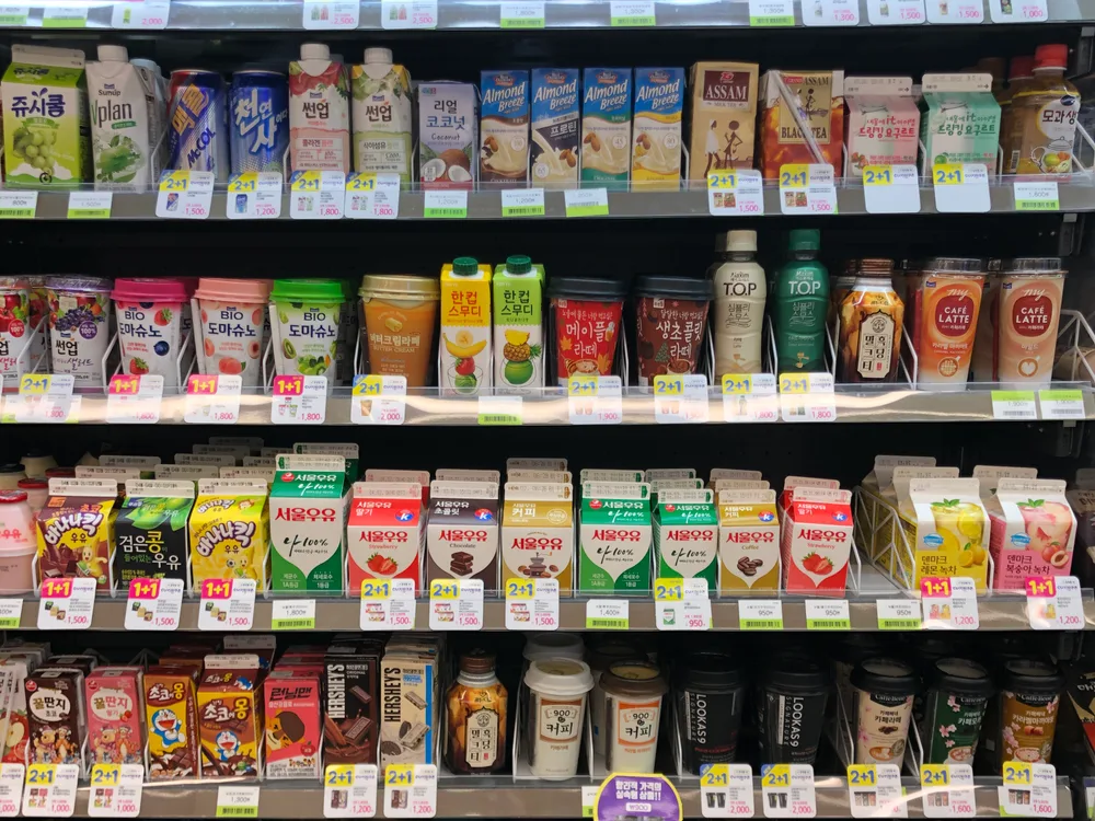 A packed convenience-store drinks fridge of milks, bottled coffees and teas, with many yellow and pink 1+1 and 2+1 promotion tags on the shelf edges