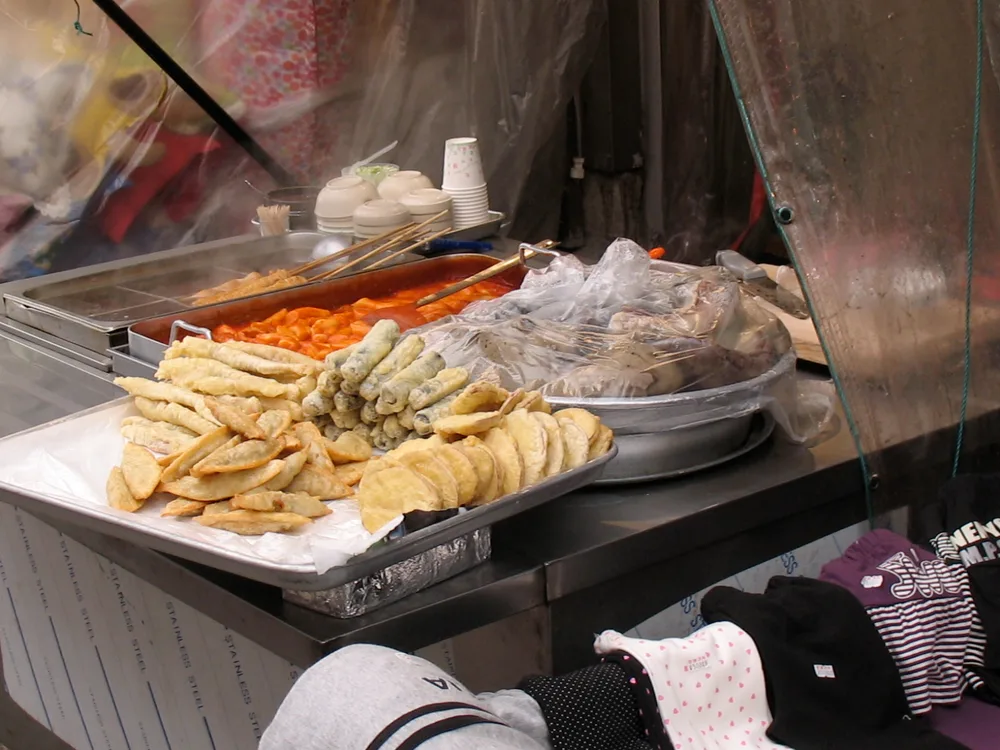 A bunsik stall with a tray of assorted twigim fritters, a bubbling trough of red tteokbokki, eomuk skewers in broth, and sundae under plastic