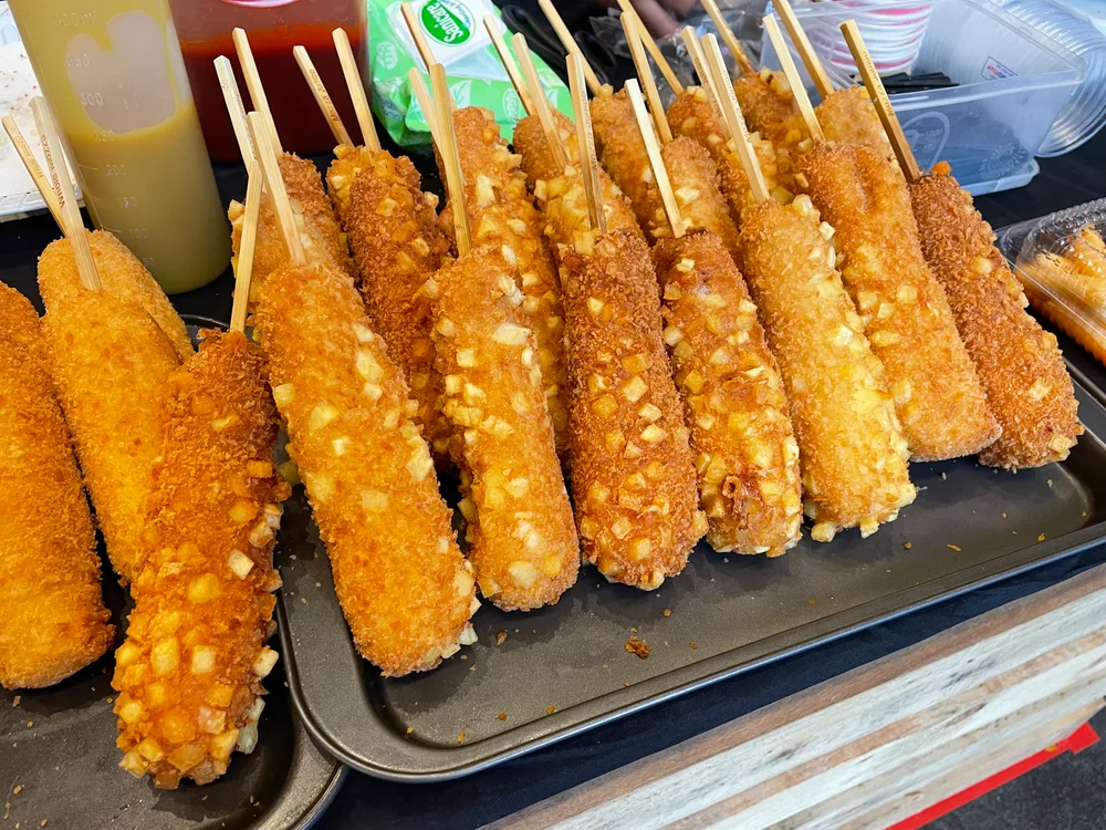 A tray of Korean corn dogs on wooden sticks, some crumb-coated and some studded with cubed potato, with squeeze bottles of sauce behind