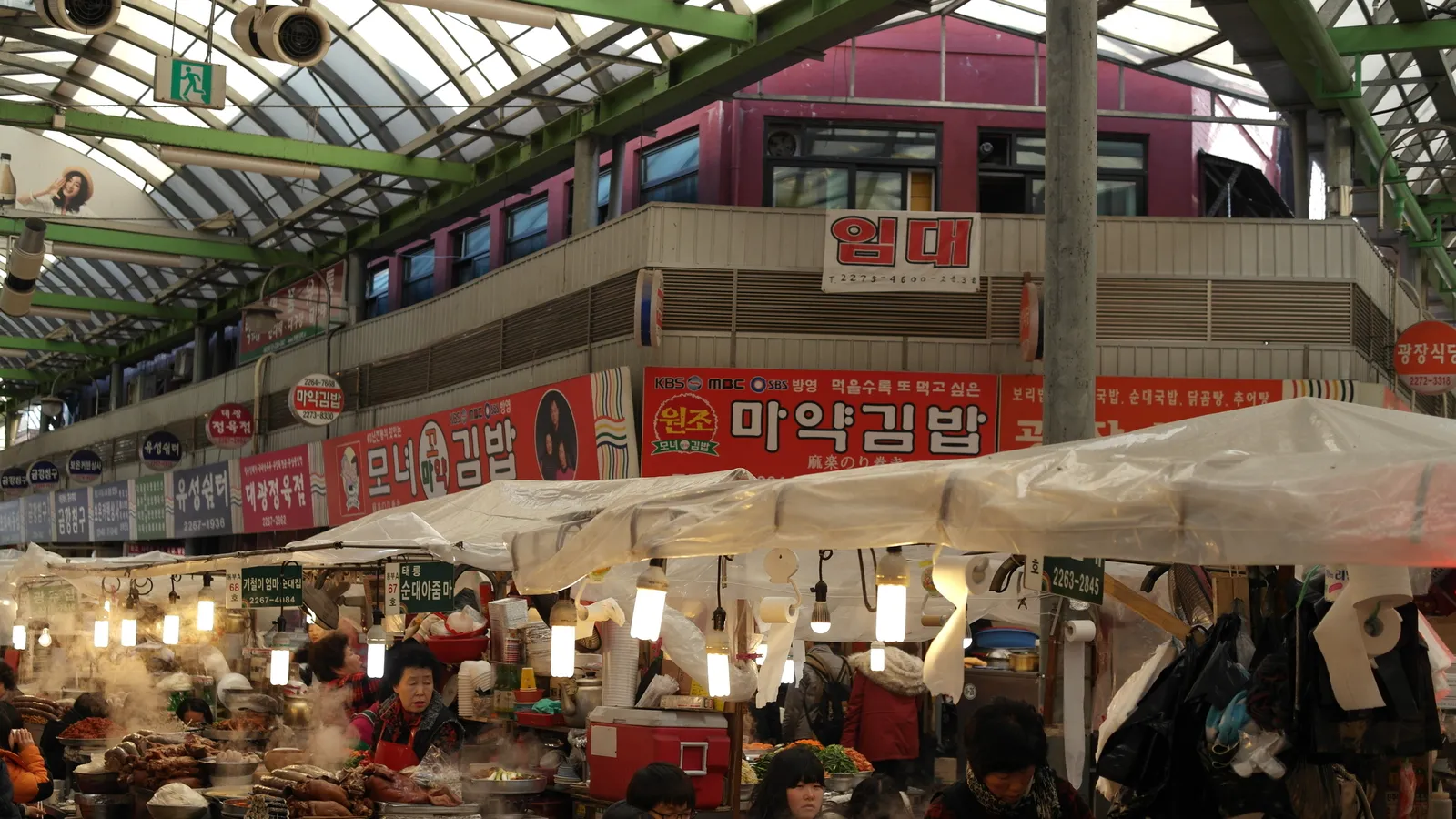 The covered food alley of Gwangjang Market in Seoul, lined with tented stalls, steaming pots and hanging signs, with people eating at the counters