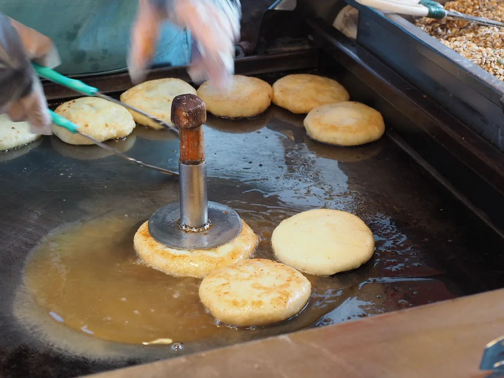 A vendor pressing rounds of hotteok dough flat on an oiled griddle with a metal press, a tray of seeds and nuts alongside