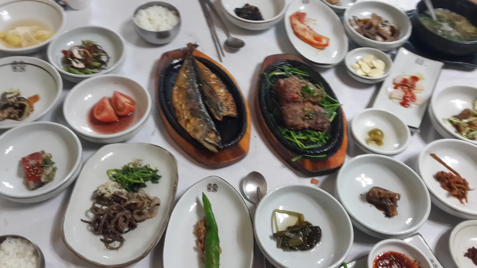 A restaurant table crowded with a hanjeongsik spread — dozens of small dishes of banchan, grilled fish, sizzling bulgogi, rice bowls and stew