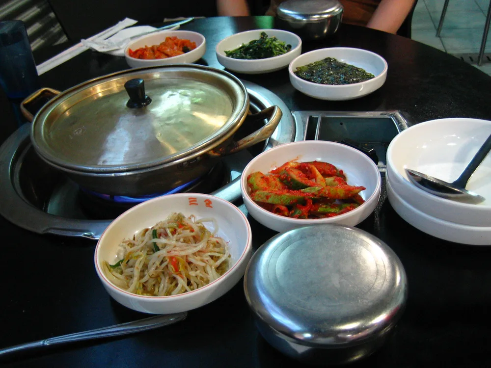 A restaurant table with a covered stew pot on a burner, lidded metal rice bowls, and five small dishes of banchan around it